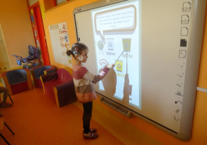 Dziewczynka stoi pod tablicą interaktywną i wskaźnikiem pokazuje kosz na odpady szklane.